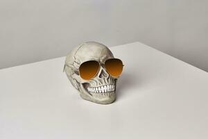 Realistic model of a human skull with teeth in a yellow sunglasses on a light table, white background. Medical science or Halloween horror concept. photo