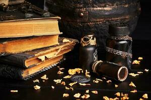 Books with spells, old pot, jars of potion are on a wooden dark table. Petals of dried roses are scattered nearby. Black background. Close-up shot. photo