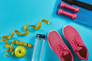 Sneakers with measuring tape on cyan blue background. Centimeter in yellow color, pink sneakers, dumbbells and bottle of water, copy space. photo