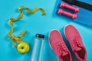 Sneakers with measuring tape on cyan blue background. Centimeter in yellow color, pink sneakers, dumbbells and bottle of water, copy space. photo