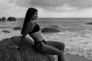 Beautiful brunette woman  in black swimwear posing on  rocky beach.  Looking on waves. Black and white. photo