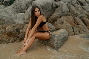 Seductive brunette woman with perfect figure posing on tropical rocky beach. Wearing stylish black swimwear. photo