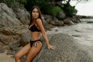 hermosa morena mujer en negro trajes de baño posando en rocoso playa. foto