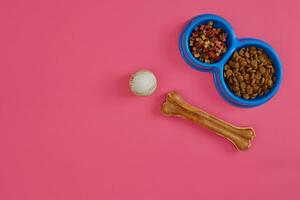 Dry pet food in bowl with a ball and dog bone on pink background top view photo