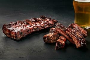 Pork ribs in barbecue sauce and a glass of beer on a black slate dish. A great snack to beer on a dark stone background. Top view with copy space photo