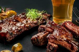 Cerdo costillas en parilla salsa y un vaso de cerveza en un negro pizarra plato. un genial bocadillo a cerveza en un oscuro Roca antecedentes. parte superior ver con Copiar espacio foto