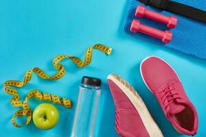 Sneakers with measuring tape on cyan blue background. Centimeter in yellow color, pink sneakers, dumbbells and bottle of water, copy space. photo