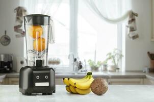 Metal food blender close-up with fresh exotic tropic fruits next to it on kitchen background with empty space. Sun flare photo