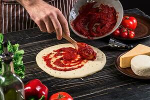 un masculino mano extensión tomate puré en un Pizza base con cuchara en un antiguo de madera antecedentes foto