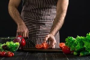 Man preparing salad on a wooden table. Men's hands cut the tomato to make a salad on black background photo