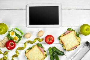 blanco tableta computadora con un blanco pantalla en el cocina mesa con sándwiches, Fresco verduras, botella de agua y manzana. almuerzo foto