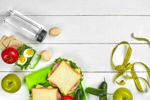 Lunch boxes with sandwiches and fresh vegetables, bottle of water and eggs on white wooden background. Top view with copy space photo