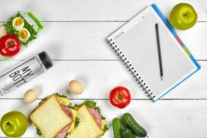 Top view planning notebook with copy space and healthy breakfast. Sandwich with ham and cheese, eggs, cucumber, tomato and bottle of water on white wooden table. photo