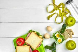 Lunch boxes with sandwiches and fresh vegetables, bottle of water and eggs on white wooden background. Top view with copy space photo