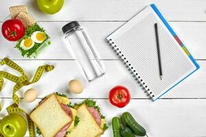 Top view planning notebook with copy space and healthy breakfast. Sandwich with ham and cheese, eggs, cucumber, tomato and bottle of water on white wooden table. photo