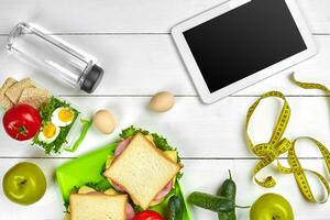 Sandwich, fresh vegetables, bottle of water and eggs on a table with a tablet, top view. Copy space photo