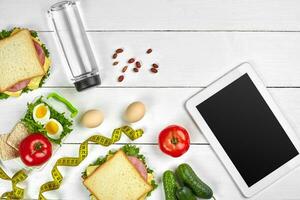 White tablet computer with a blank screen on the kitchen table with sandwiches, fresh vegetables, bottle of water and apple. Lunch photo