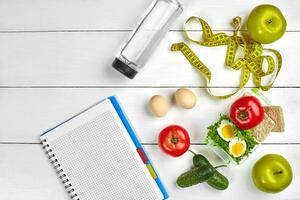 Top view planning notebook with copy space and healthy breakfast. Sandwich with ham and cheese, eggs, cucumber, tomato and bottle of water on white wooden table. photo