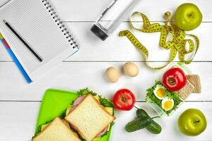 Top view planning notebook with copy space and healthy breakfast. Sandwich with ham and cheese, eggs, cucumber, tomato and bottle of water on white wooden table. photo