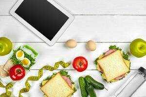 blanco tableta computadora con un blanco pantalla en el cocina mesa con sándwiches, Fresco verduras, botella de agua y manzana. almuerzo foto