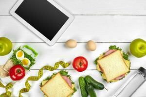 White tablet computer with a blank screen on the kitchen table with sandwiches, fresh vegetables, bottle of water and apple. Lunch photo