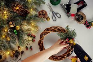 Top view of female hands make a Christmas wreath. Packed gifts and scrolls, spruce branches and tools on white table. photo