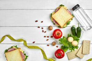 Lunch boxes with sandwiches and fresh vegetables, bottle of water, nuts and eggs on white wooden background. Top view with copy space photo