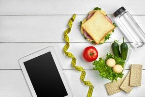 Sandwich, fresh vegetables, bottle of water and eggs on a table with a tablet, top view. Copy space photo