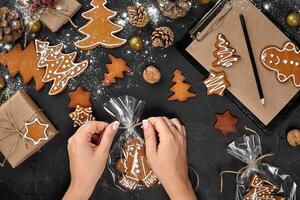 Christmas gift gingerbread on dark background. Biscuits in festive packaging. Woman is packaging Christmas gingerbread cookies with icing sugar. Top view photo