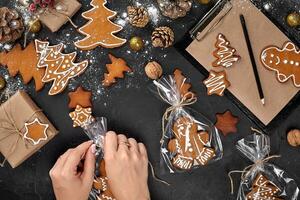 Christmas gift gingerbread on dark background. Biscuits in festive packaging. Woman is packaging Christmas gingerbread cookies with icing sugar. Top view photo