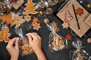 Christmas gift gingerbread on dark background. Biscuits in festive packaging. Woman is packaging Christmas gingerbread cookies with icing sugar. Top view photo