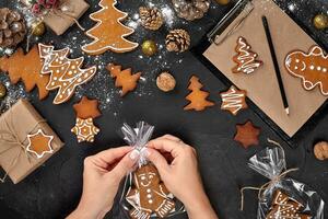 Christmas gift gingerbread on dark background. Biscuits in festive packaging. Woman is packaging Christmas gingerbread cookies with icing sugar. Top view photo