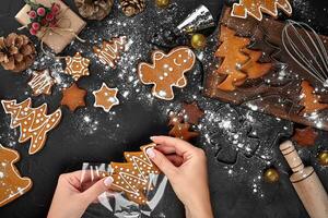 Christmas gift gingerbread on dark background. Biscuits in festive packaging. Woman is packaging Christmas gingerbread cookies with icing sugar. Top view photo