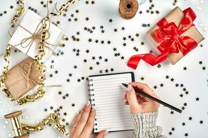 Festive decorations, delicate wavy ribbons, metallic star shaped confetti and notebook with wish list on white table, flat lay style. Christmas holidays decoration concept. photo