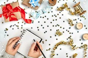 Festive decorations, delicate wavy ribbons, metallic star shaped confetti and notebook with wish list on white table, flat lay style. Christmas holidays decoration concept. photo