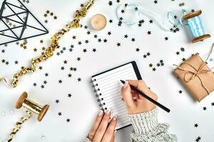 Festive decorations, delicate wavy ribbons, metallic star shaped confetti and notebook with wish list on white table, flat lay style. Christmas holidays decoration concept. photo