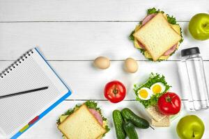 Top view planning notebook with copy space and healthy breakfast. Sandwich with ham and cheese, eggs, cucumber, tomato and bottle of water on white wooden table. photo