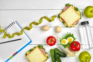 Top view planning notebook with copy space and healthy breakfast. Sandwich with ham and cheese, eggs, cucumber, tomato and bottle of water on white wooden table. photo