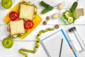 Top view planning notebook with copy space and healthy breakfast. Sandwich with ham and cheese, eggs, cucumber, tomato and bottle of water on white wooden table. photo