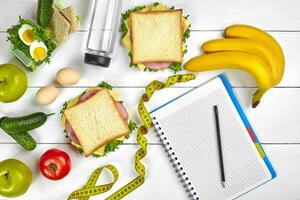 Top view planning notebook with copy space and healthy breakfast. Sandwich with ham and cheese, eggs, cucumber, tomato and bottle of water on white wooden table. photo