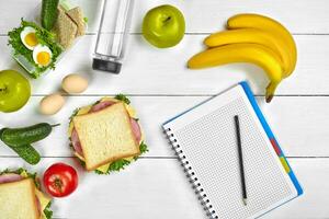 Top view planning notebook with copy space and healthy breakfast. Sandwich with ham and cheese, eggs, cucumber, tomato and bottle of water on white wooden table. photo