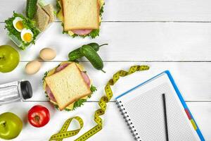 Top view planning notebook with copy space and healthy breakfast. Sandwich with ham and cheese, eggs, cucumber, tomato and bottle of water on white wooden table. photo