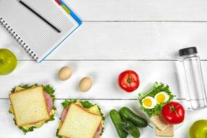 Top view planning notebook with copy space and healthy breakfast. Sandwich with ham and cheese, eggs, cucumber, tomato and bottle of water on white wooden table. photo