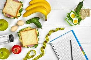 Top view planning notebook with copy space and healthy breakfast. Sandwich with ham and cheese, eggs, cucumber, tomato and bottle of water on white wooden table. photo