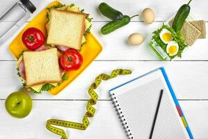 Top view planning notebook with copy space and healthy breakfast. Sandwich with ham and cheese, eggs, cucumber, tomato and bottle of water on white wooden table. photo