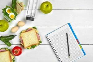 Top view planning notebook with copy space and healthy breakfast. Sandwich with ham and cheese, eggs, cucumber, tomato and bottle of water on white wooden table. photo