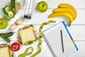 Top view planning notebook with copy space and healthy breakfast. Sandwich with ham and cheese, eggs, cucumber, tomato and bottle of water on white wooden table. photo