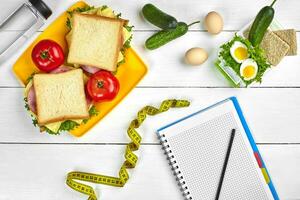 Top view planning notebook with copy space and healthy breakfast. Sandwich with ham and cheese, eggs, cucumber, tomato and bottle of water on white wooden table. photo