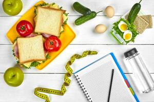 Top view planning notebook with copy space and healthy breakfast. Sandwich with ham and cheese, eggs, cucumber, tomato and bottle of water on white wooden table. photo