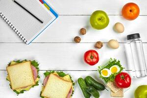 Top view planning notebook with copy space and healthy breakfast. Sandwich with ham and cheese, eggs, cucumber, tomato and bottle of water on white wooden table. photo